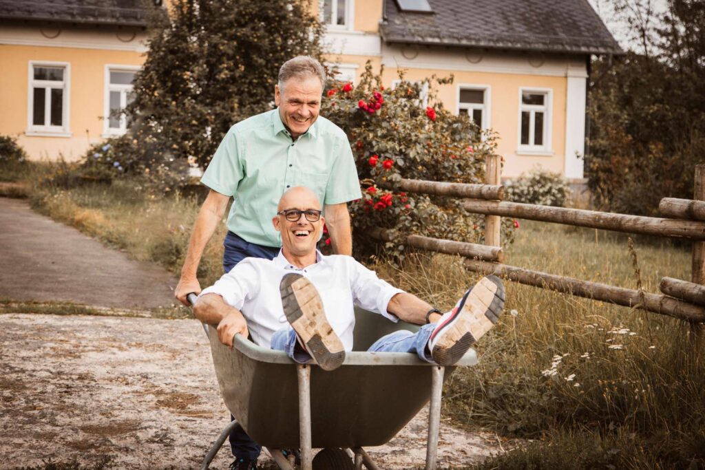 Zwei Maenner, einer sitzend in einer Schubkarre und der andere schiebt ihn im Garten.